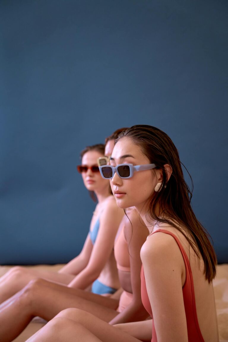 Three diverse women pose in swimsuits and sunglasses against a blue background.