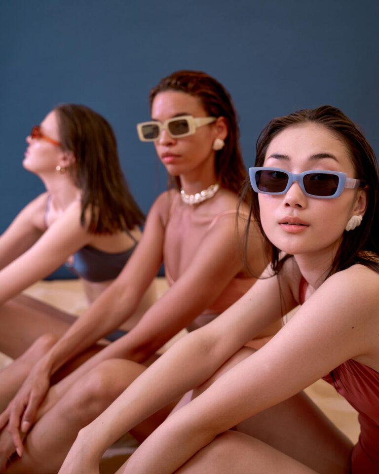 Three women sitting indoors in stylish swimwear and sunglasses, exuding modern fashion vibes.